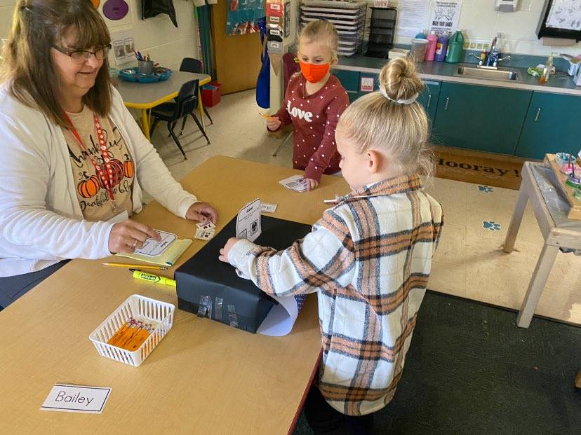 Preschool students places ballot in box as teacher and other student watch